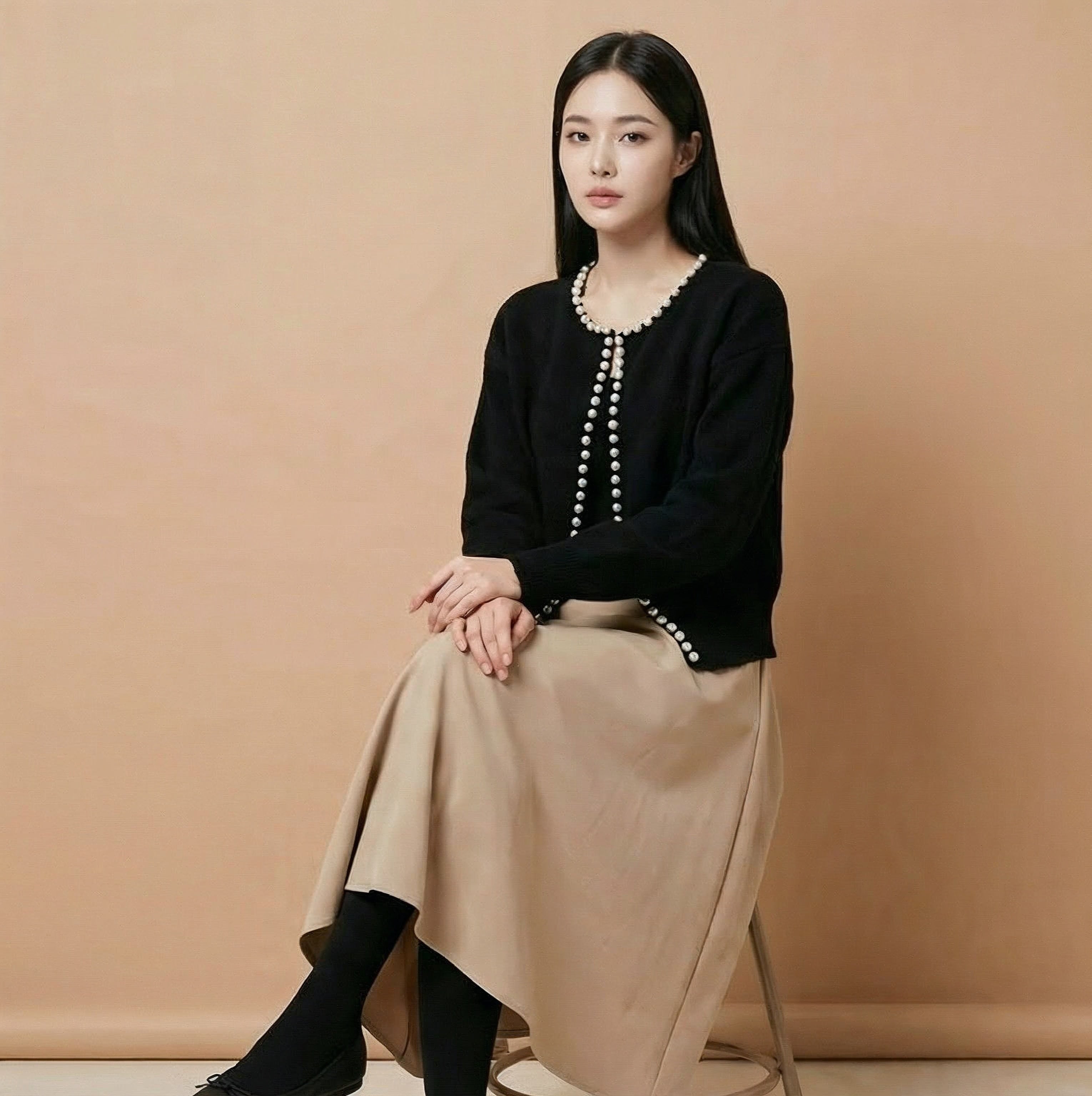Cropped v neck cardigan in black worn by a seated model, showing a short length, long sleeves, pearl button detailing, and open-front finish