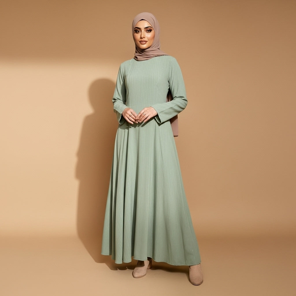 Long sleeve winter maxi dress in sage green worn by a standing hijabi model facing front, showing a round neckline, full-length sleeves, softly textured fabric, relaxed A-line silhouette, floor-grazing hem, and smooth vertical drape