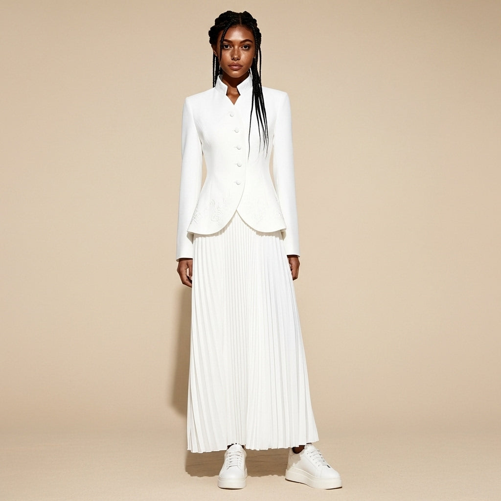 Skirt suit set in white worn by a standing model, showing a tailored stand-collar jacket with button closure, sculpted hem shape, and a pleated maxi skirt with full coverage