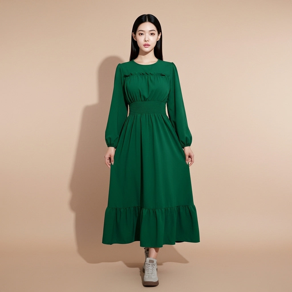 Shirred waist maxi dress in deep green worn by a standing model facing front, showing a round neckline, long gathered sleeves, elasticated shirred waist panel, smooth draped fabric through the skirt, full-length silhouette, and softly flared lotus hem