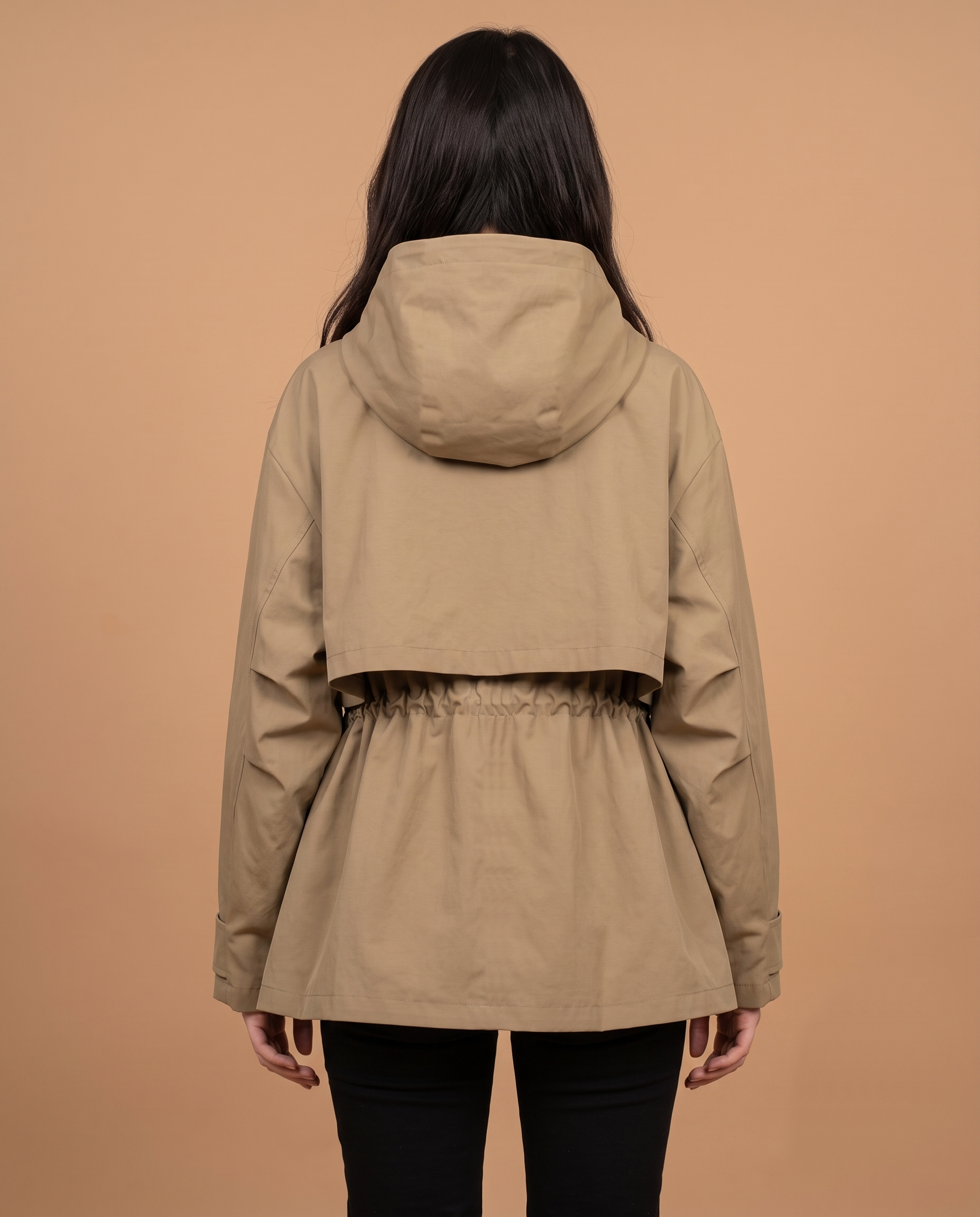 Utility jacket in khaki worn by a standing female model facing back, showing a hooded back panel, gathered waist detail, long sleeves with cuff tabs, mid-thigh length hemline, and relaxed back drape over trousers