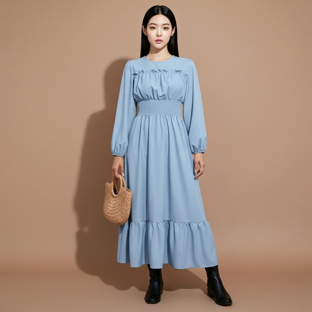 Shirred waist maxi dress in light blue worn by a standing model facing front, showing a round neckline, long sleeves with elastic cuffs, textured shirred waist, fluid full-length skirt, softly pleated lotus hem, and straight vertical drape to the ankles